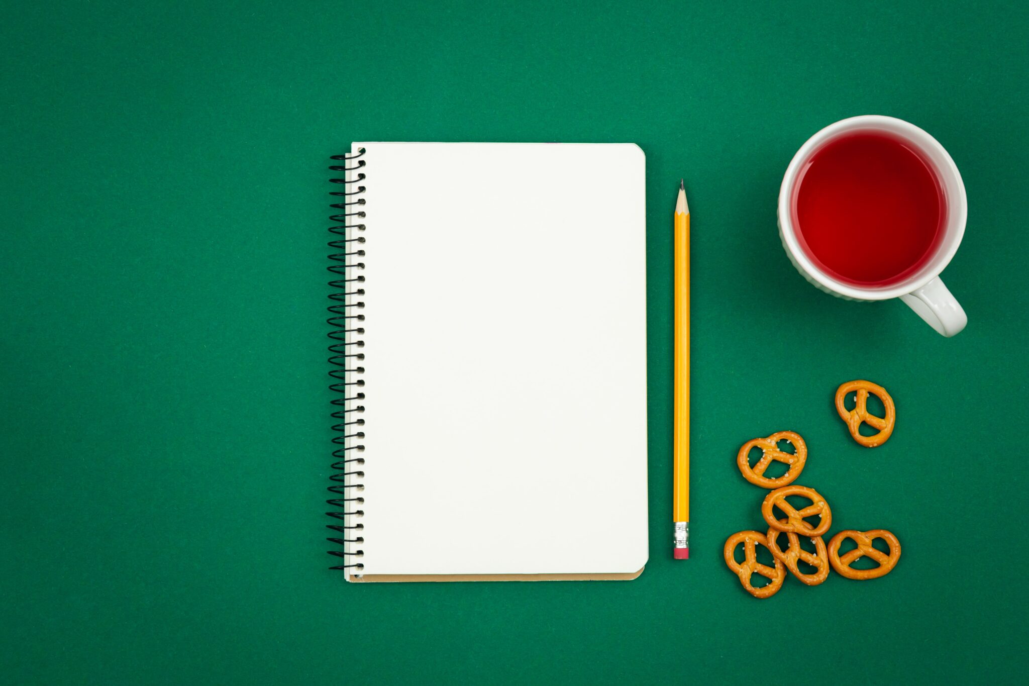 Notizbuch, Bleistift, Tee und Brezeln auf grünem Tisch – optimale Voraussetzungen für kreative Schreibsessions.